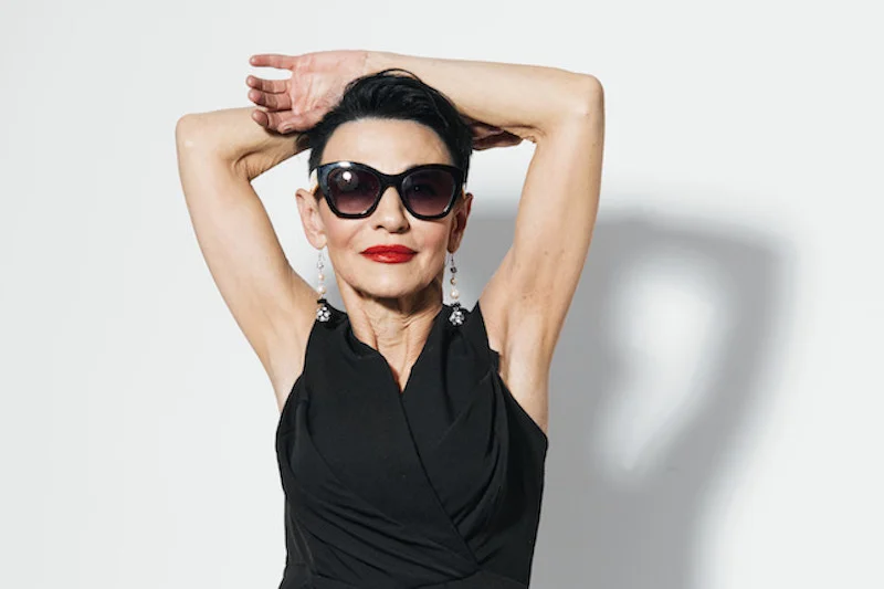 Une femme senior aux cheveux noirs courts, des lunettes de soleil et un rouge à lèvres rouge - les bras relevés - un top sans manches noir